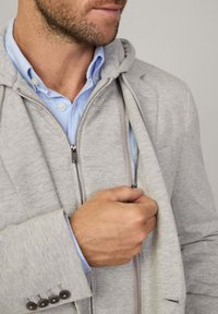 Grå zip-up huvtröja som är lager på lager över en ljusblå skjorta med knappar, med mjuk textur, synliga dragkedjedetaljer och en stukturerad krage.