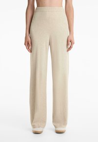 Beige, hoch taillierte, weit geschnittene Strickhose, getragen mit Schuhen mit weißer Sohle, vom Taillenbereich bis zu den Füßen auf einem weißen Hintergrund gezeigt.