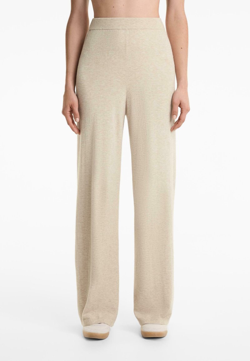 Beige, hoch taillierte, weit geschnittene Strickhose, getragen mit Schuhen mit weißer Sohle, vom Taillenbereich bis zu den Füßen auf einem weißen Hintergrund gezeigt.