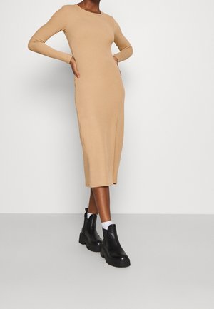 Beige Midi-Kleid mit langen Ärmeln und taillierter Silhouette. Das Model trägt schwarze Stiefeletten und weiße Socken, vor einem schlichten Hintergrund.
