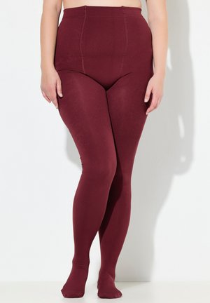 Person, der burgunderfarbene, hochtaillierte, lange Leggings trägt, steht entspannt mit den Händen an den Seiten gegen eine weiße Wand.