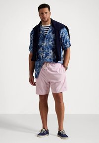 Homem com uma camisa de flores azul de mangas curtas, uma camisola riscada por baixo, calções rosas, sapatos azul-marinho e um suéter azul-marinho colocado sobre os ombros, em pé contra um fundo branco.