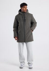Giacca isolante verde oliva con cappuccio, chiusura frontale con zip e due tasche con zip. Indossata sopra pantaloni della tuta grigi chiari e sneakers bianche.