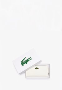 Portefeuille blanc avec un logo de crocodile vert, affichant une texture lisse et une fermeture éclair, présenté dans une boîte blanche avec un logo assorti.