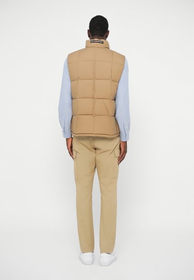 Gilet imbottito beige con design a trapunta, dotato di un colletto alto e una toppa con logo, indossato sopra una camicia azzurro chiaro, abbinato a pantaloni cargo khaki.