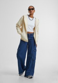Crème ritsvak hoodie over een witte cropped t-shirt, gecombineerd met wijde blauwe denim cargobroek met zakken en rafelige zoom.