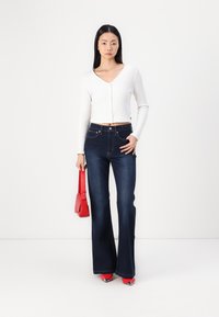 Top lungo bianco a coste a maniche lunghe con chiusura a bottoni, abbinato a jeans svasati blu scuro. La modella tiene in mano una piccola borsa rossa e indossa tacchi rossi.