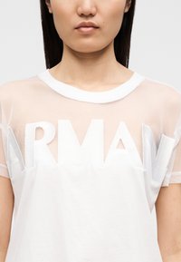 Λευκό t-shirt με διαφανείς πλεκτές μανίκια και μεγάλο κεντημένο λογότυπο "ARMANI" στο μπροστινό μέρος. Μαλακό ύφασμα με άνετη εφαρμογή.