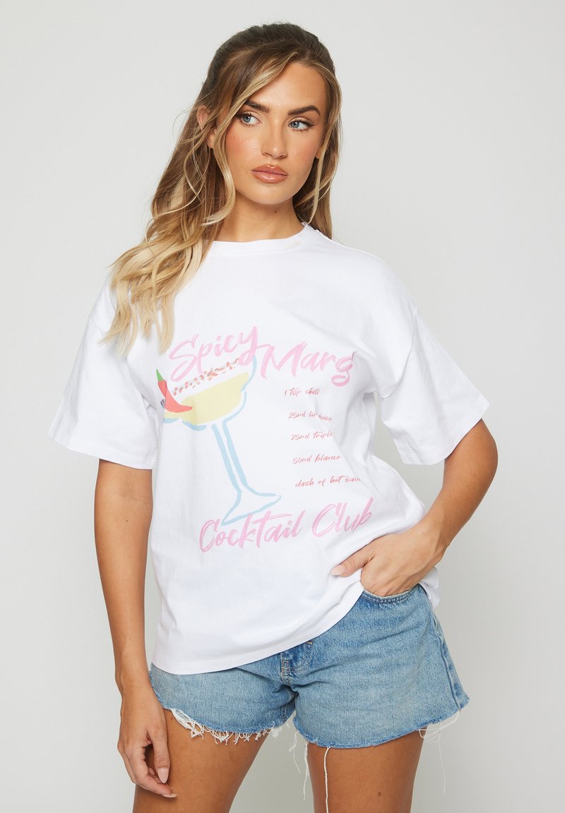 Kvinna med långt blont hår som bär en vit t-shirt med en illustration av Spicy Marg Cocktail Club och jeansshorts, poserande med handen i fickan.