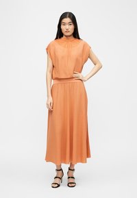 Kvinde stående med hånden på hoften iført en ærmeløs orange midikjole og sorte ankelstrop-hæle mod en hvid baggrund.