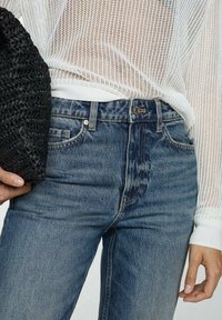 Jeans in denim blu con effetto sbiadito, caratterizzati da un classico design a cinque tasche e chiusura con bottone in metallo. Indossati con un top in maglia bianca e una borsa nera.