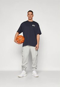 Camiseta extragrande azul marino con logo, combinada con pantalones cargo deportivos gris claro y zapatillas blancas. El modelo sostiene un balón de baloncesto naranja.