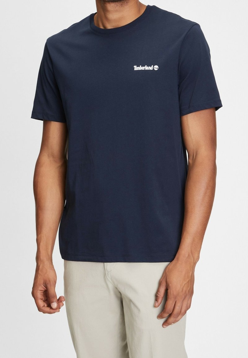 Homme portant un t-shirt Timberland bleu marine à manches courtes et un pantalon beige clair, debout devant un fond blanc uni.