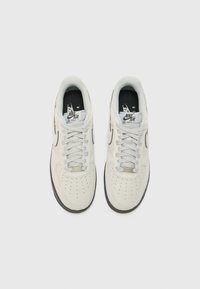 White suede sneakers with round toe, perforated details, flat laces, and black accents. Features prominent branding on tongue and sides.