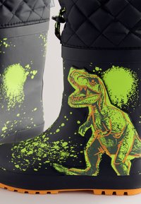 Stivali da pioggia in gomma nera con una vivace grafica di un dinosauro verde e motivi a schizzo, con suola arancione e design superiore trapuntato.
