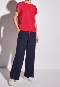 Röd kortärmad topp med dragsko vid fållen, tillsammans med mörkblå vida byxor och vita sneakers, mot en ljus bakgrund.