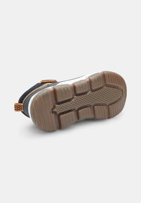 Zapatilla en blanco y negro con una suela de goma texturizada, detalles en naranja y un diseño robusto. Presenta un collar acolchado y una superficie de agarre texturizada.