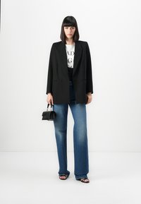 Zwarte oversized blazer, wit graphic T-shirt, high-waisted blauwe denim jeans, zwarte handtas en zwarte sandalen met bandjes. Effen achtergrond.