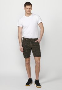 Grüne Camouflage-Shorts mit elastischem Bund und Kordelzug, ausgestattet mit Seitentaschen. Kombiniert mit einem schlichten weißen T-Shirt mit Rundhalsausschnitt und schwarzen Sneakers.