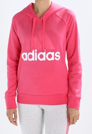 Person iført en knaldrød Adidas hættetrøje med hvidt logo og lysegrå bukser med pink striber står foran en ensfarvet baggrund.