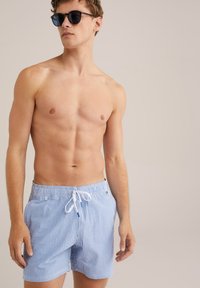 Kék és fehér csíkos fürdőnadrág, húzózsinórral ellátott derékkal. Simább anyag, laza fazon és oldalsó zsebek. Sötét napszemüveggel viselve.