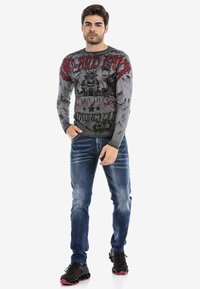 Graues Langarmshirt mit grafischem Design in Rot und Schwarz. Kombiniert mit blauen Jeans und schwarzen Sneakers mit roten Akzenten.