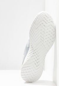 Zapatilla gris con suela de goma blanca texturizada, con un patrón en zigzag. Parte superior hecha de material transpirable con un branding mínimo.