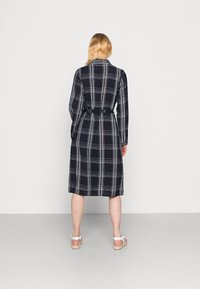 Vestido às riscas azul-marinho com mangas compridas e cintura cingida, apresentando padrões quadriculados em branco e cinza. Comprimento até metade da canela, textura de tecido suave.