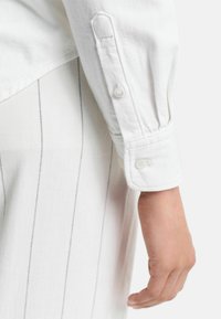 Weißes Langarmhemd mit geknöpften Manschetten, das eine strukturierte Oberfläche und subtile vertikale Streifen auf dem benachbarten Kleidungsstück aufweist.