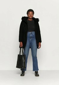 Mlad človek oblečen v črno krzneno jakno s kapuco, črno majico, modre kavbojke, črne škornje, drži črno torbo, stoji ob enakomernem ozadju.