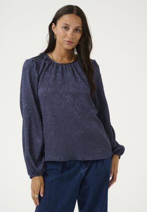 Navy blue blouse with textured fabric, long puffed sleeves, gathered neckline, and a loose fit; paired with dark blue jeans.