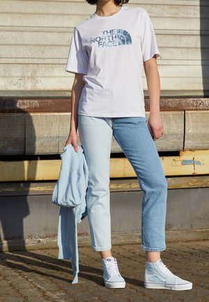 Witte t-shirt met blauwe grafische logo, lichtblauwe spijkerbroek en lichtblauwe hoge sneakers. Een bijpassende lichtblauwe sweatshirt hangt over de arm.