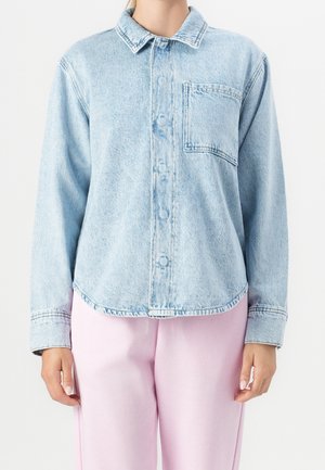 Persona que lleva una camisa vaquera azul claro con botones y un bolsillo en el pecho, y pantalones rosa claro, de pie con los brazos relajados a los lados.