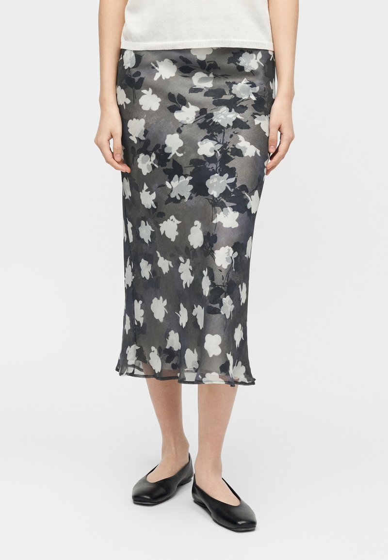 Μαύρη και άσπρη φλοράλ midi φούστα φορεμένη με μαύρα φλατ παπούτσια και λευκό τοπ, εμφανιζόμενη από τη μέση έως τα πόδια σε ουδέτερο φόντο.