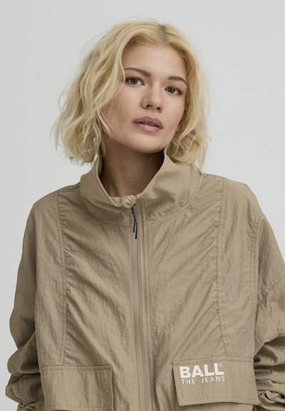 Veste beige avec col montant, fermeture éclair et deux poches. Présente un tissu texturé et un patch logo sur la poche gauche.
