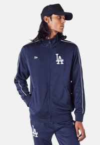 Navy Zip-Up Trainingsjacke aus glattem Stoff, mit weißen Streifen an den Ärmeln und einem weißen "LA"-Logo auf der Brust.