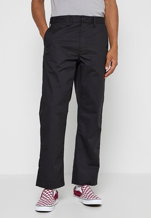 Uomo che indossa pantaloni neri a gamba dritta con le mani nelle tasche e sneakers slip-on a quadretti rossi e bianchi su sfondo semplice.