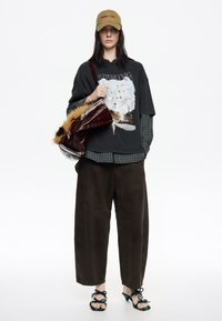 Camiseta gráfica negra de gran tamaño superpuesta sobre una camisa de cuadros verde, combinada con pantalones de pierna ancha marrones y sandalias negras de tacón con lazos decorativos.