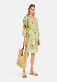 Blumendruckkleid mit drei Viertelärmeln in Hellgrün, Gelb und Pink, kombiniert mit einer geflochtenen Strohtasche und beigen Slides.