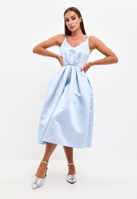 Abito in satin blu chiaro con scollatura a V, corpetto aderente e gonna ampia con pieghe. Abbinato a tacchi a punta argentati con dettagli a fiocco.