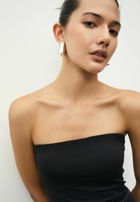 Svart strapless topp med ribbad textur. Modellen bär matte, gyllene geometriska örhängen. Hudtonen är ljus. Bakgrunden är helt vit.