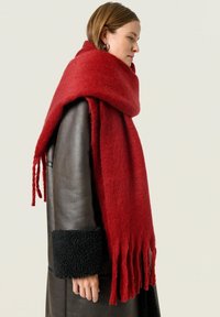Rød uldscarf med frynsede kanter, båret over en mørkebrun læderjakke med sorte shearling-manchetter. Blød tekstur og varm farve.