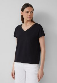 T-shirt noir à manches courtes et col en V avec une bordure en dentelle festonnée autour de l'encolure, fabriqué dans un tissu doux, porté avec un pantalon blanc.