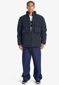 Puffjacka i mörk marinblå, har flera framfickor, hög krage, dragkedjestängning, kombinerad med lösa denimbyxor och svarta sneakers.