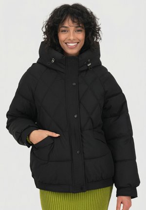 Schwarze gesteppte Steppjacke mit Kapuze, versehen mit vorderen Druckknöpfen, zwei Seitentaschen und gerippten Bündchen. Das Modell trägt einen grünen, gerippten Rock.