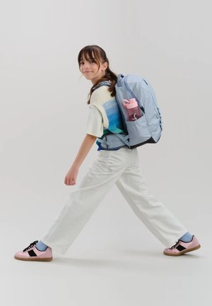 Young person walking with light blue backpack holding pink water bottle, wearing white pants, pink sneakers, and a blue-green sweater vest.