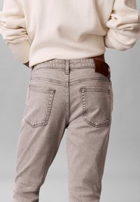 Άτομο που φοράει ανοιχτό μπεζ τζιν και εκρού πουλόβερ, εμφανιζόμενο από πίσω στο ύψος της μέσης, μπροστά σε γκρι φόντο.