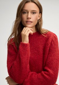 Maglione rosso lavorato a maglia con un motivo testurizzato, alto collo e maniche lunghe con dettagli a coste. Materiale morbido e spesso.