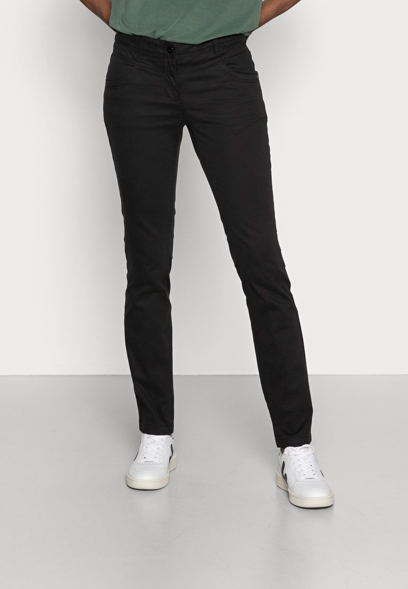 Personne portant un pantalon noir slim et des baskets blanches, debout les mains dans le dos devant un fond blanc uni.
