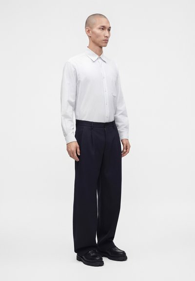 Homme debout portant une chemise blanche boutonnée, un pantalon noir à jambes larges et des chaussures noires sur un fond clair uni.
